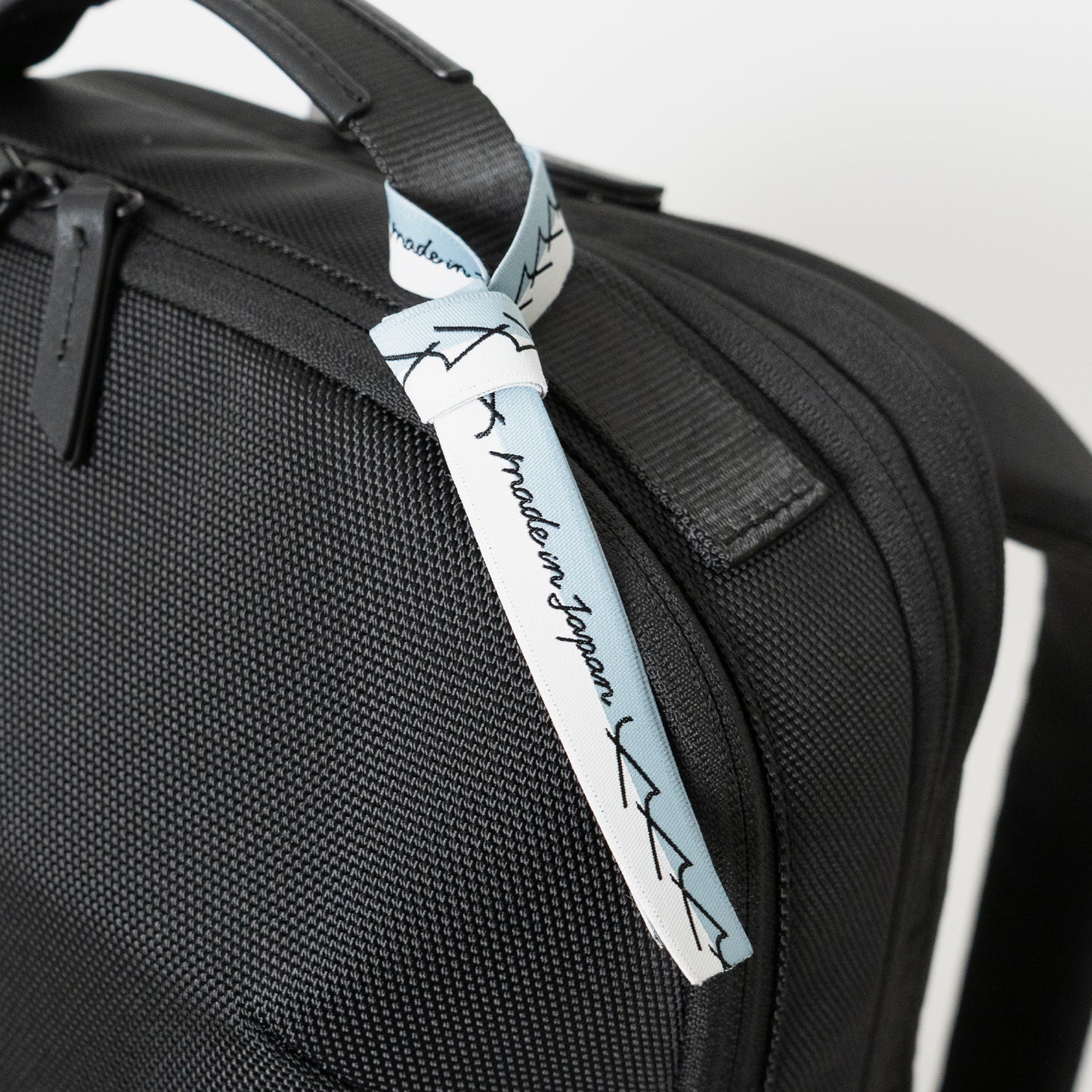 Floe ワーク＆デイリーバックパックはメイドインジャパン・日本製の証が付いています。ブランドマークの”y”を連ねたリボンのデザインで、北海道の空と雪のカラーを取り合わせました。リボンは外してお使いいただくことも可能です。